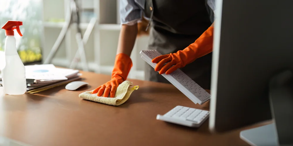 A person wearing orange rubber gloves cleans a desk with a cloth, holding a computer keyboard. A spray bottle, mouse, and computer monitor are on the desk.