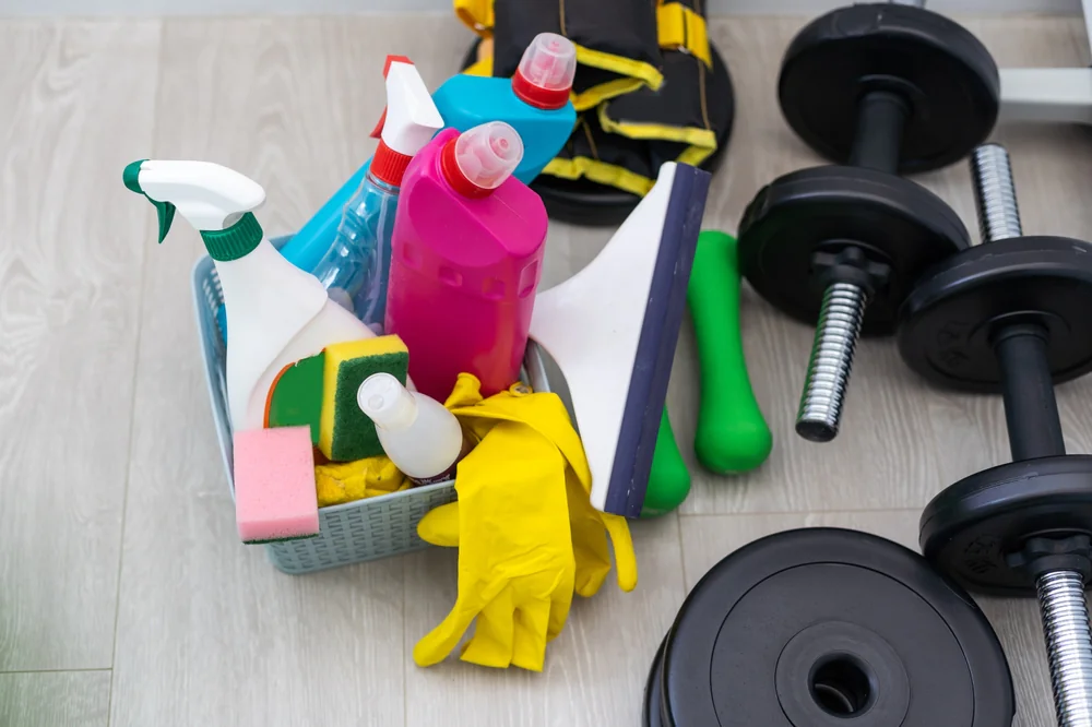 A basket filled with cleaning supplies, including spray bottles, sponges, rubber gloves, and a squeegee, sits on a light wooden floor next to black dumbbells and gym equipment.