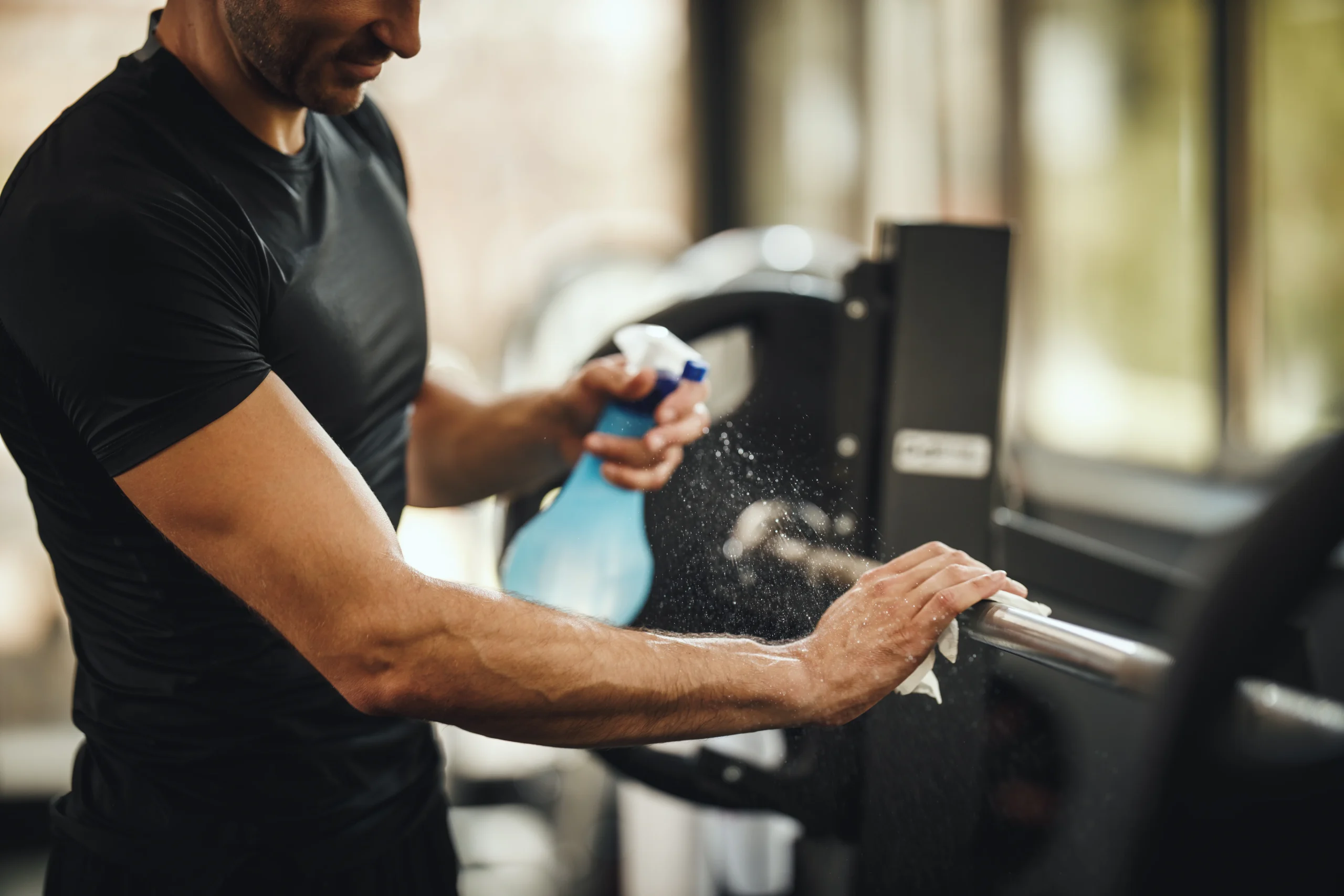 A man in a black shirt cleans a gym barbell with a cloth and spray bottle, wiping the equipment thoroughly. The gym setting is visible in the background.