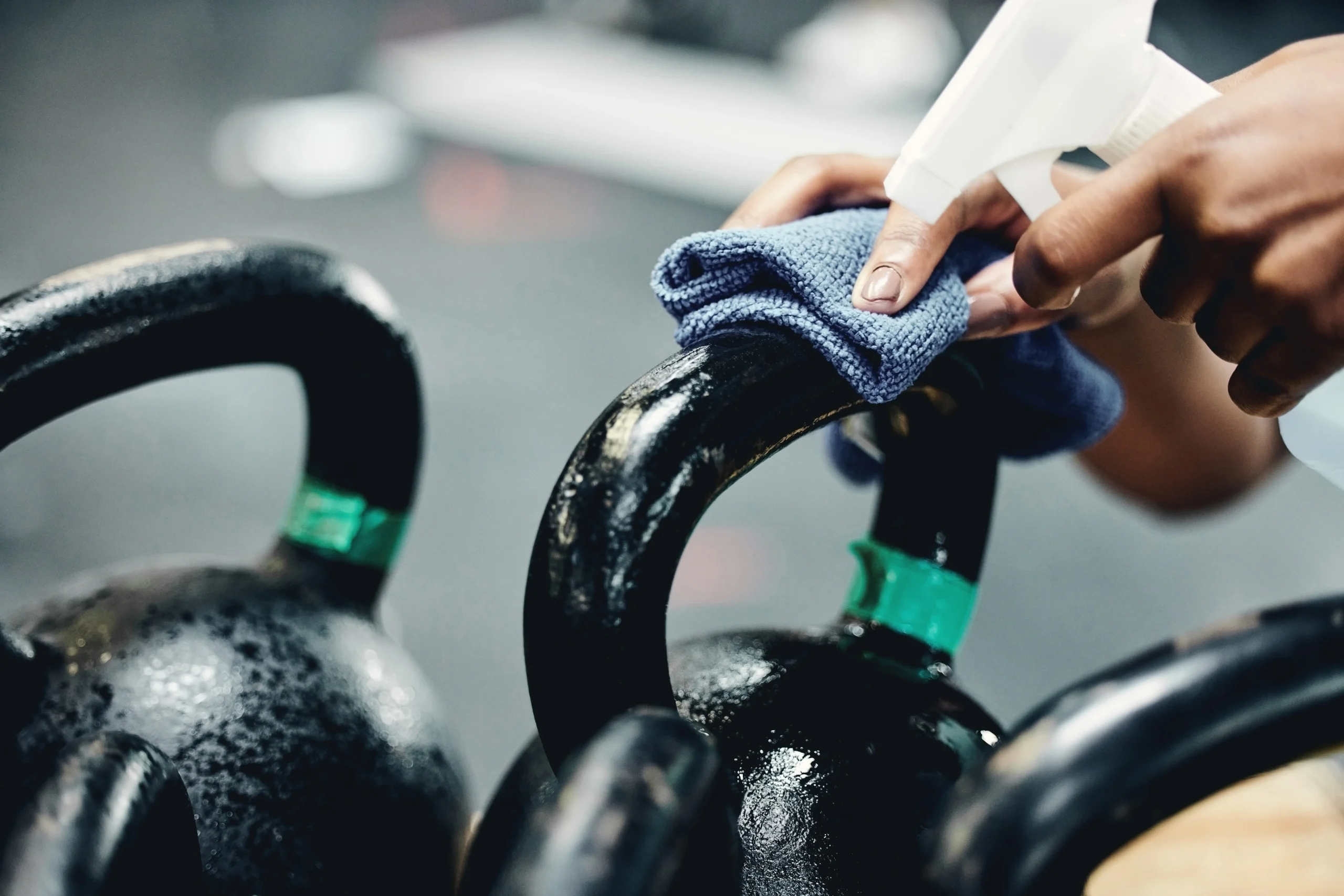 Close-up of a person cleaning a kettlebell handle with a blue cloth and spray bottle in a gym setting. Other kettlebells are visible in the background.