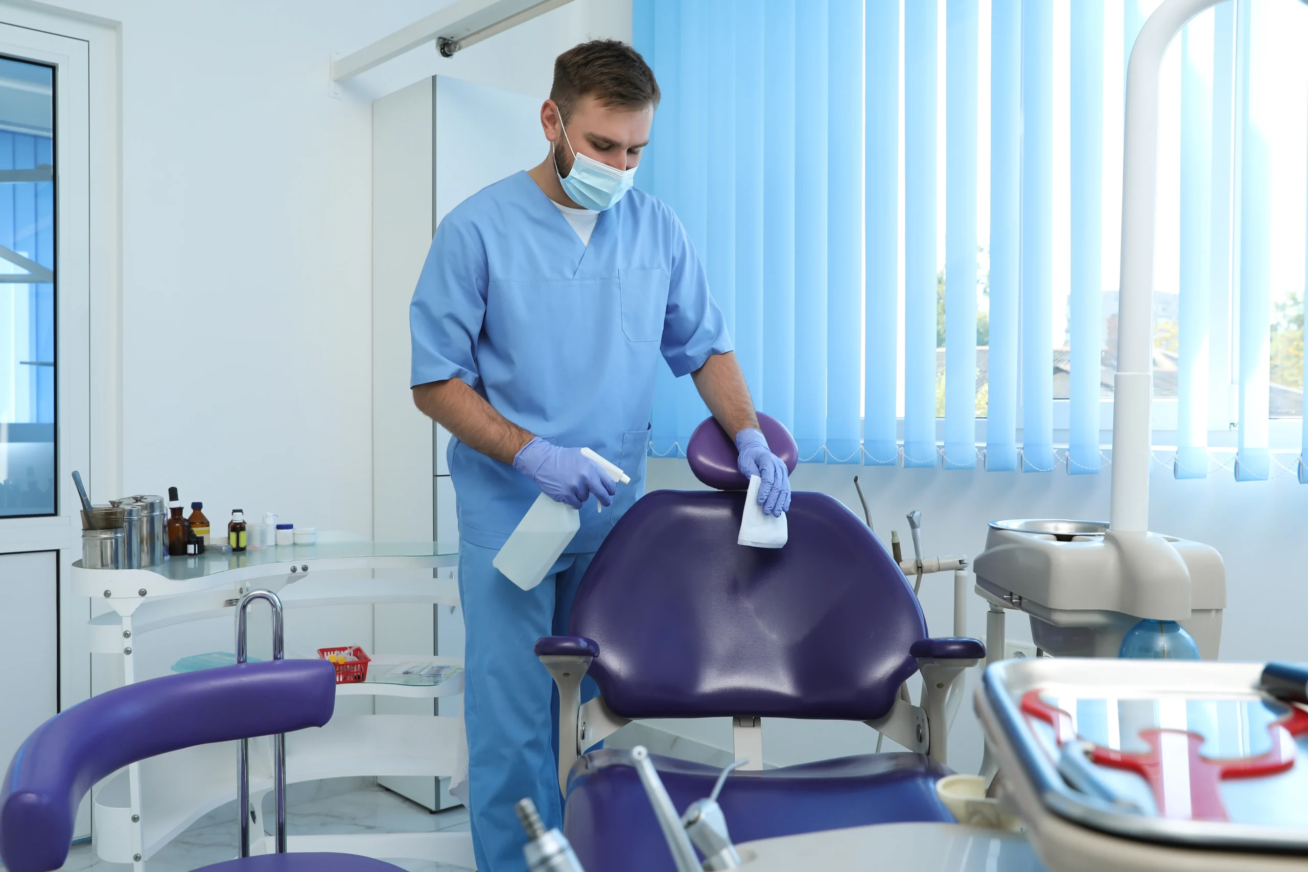 A dentist wearing scrubs, gloves, and a mask disinfects a purple dental chair with a spray bottle and cloth in a bright, modern dental clinic.