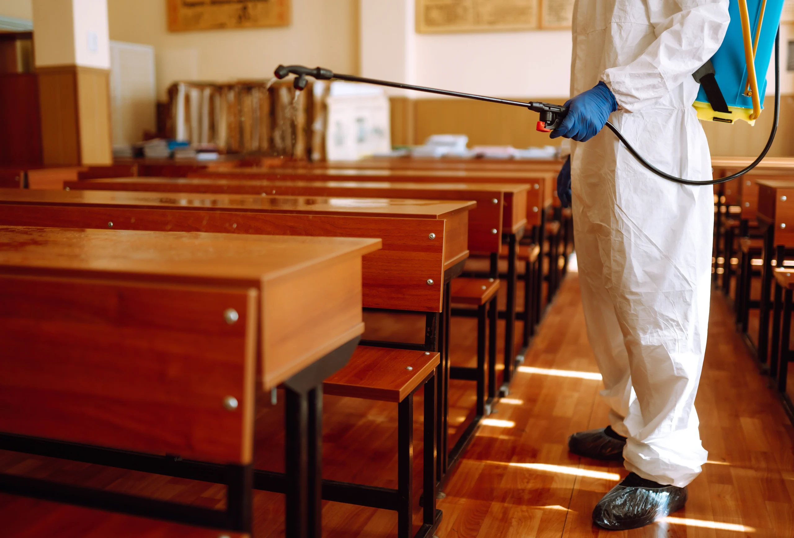 A person in protective clothing and gloves disinfects wooden desks in an empty classroom using a spray device.