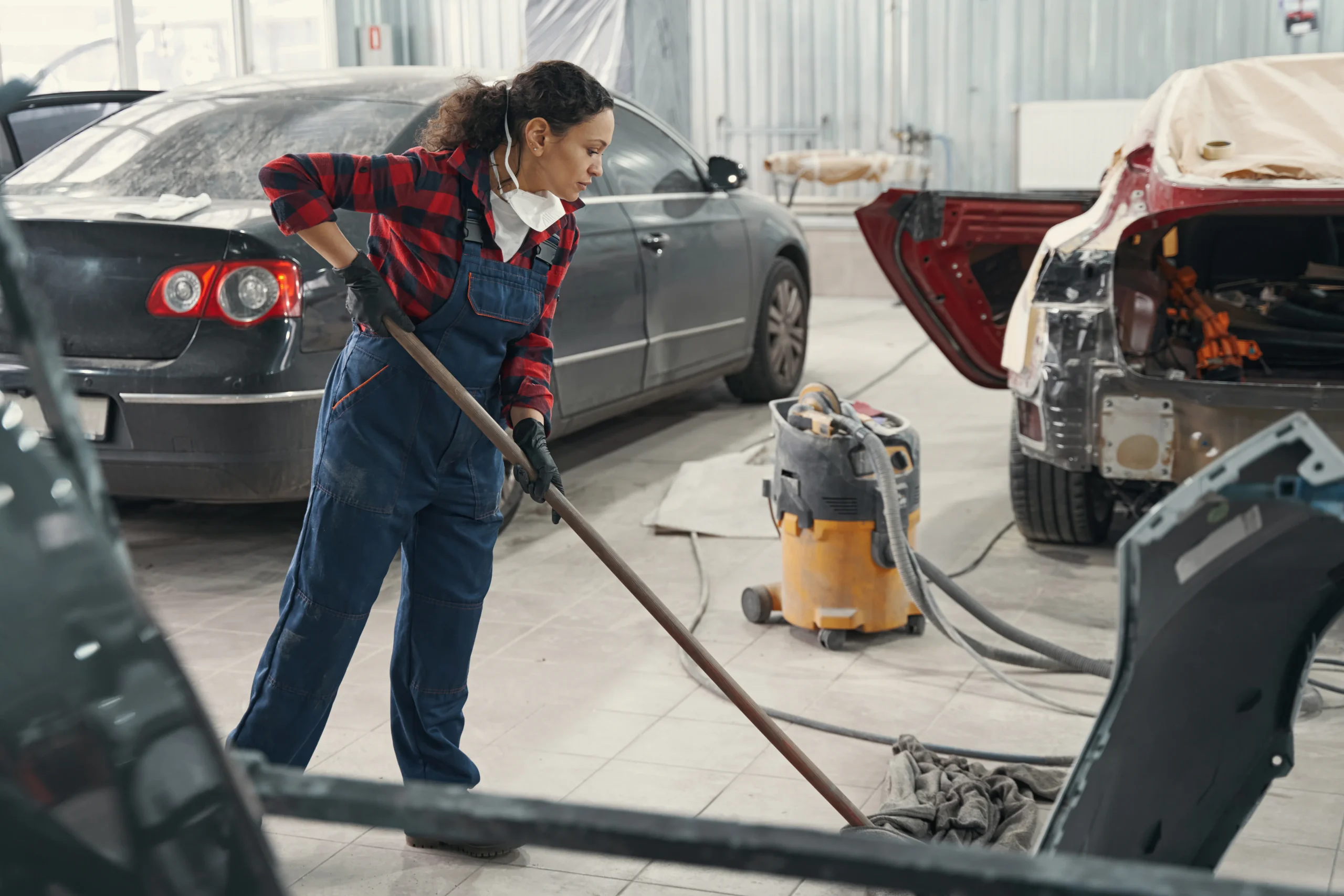 A woman in overalls and gloves sweeps the floor of an auto repair shop, surrounded by cars and repair equipment.